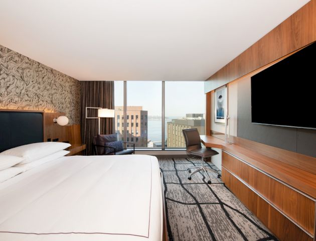 A modern hotel room with a large bed, a flat-screen TV, a desk with a chair, and a city view through floor-to-ceiling windows.