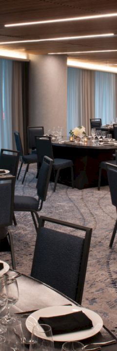 A modern, elegantly set dining room with multiple tables, black chairs, silverware, glassware, and floral arrangements, ready for a formal event.