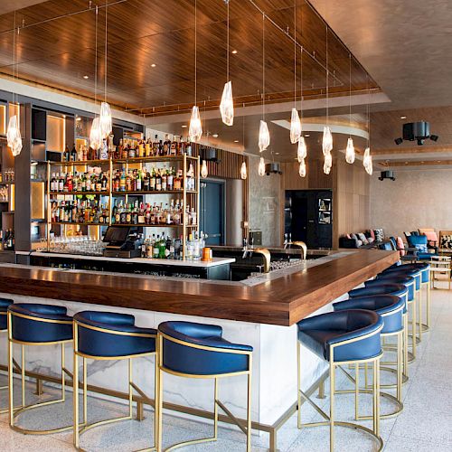 The image depicts a modern bar with a wooden counter, blue cushioned bar stools, a well-stocked bar shelf, and hanging pendant lights.