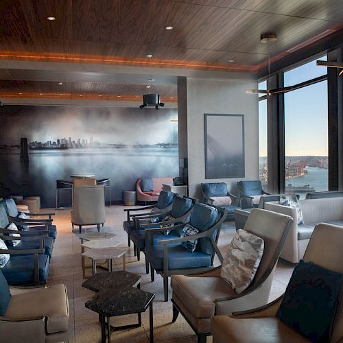 A modern lounge with rows of seating, large windows highlighting a coastal view, and contemporary decor, including wall art of a cityscape.