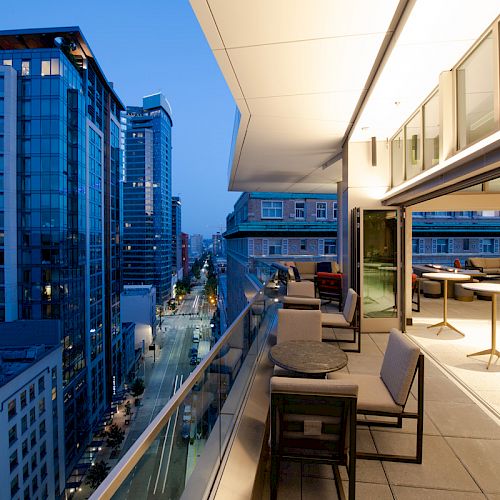 A modern balcony at dusk overlooks a cityscape with high-rise buildings and streets illuminated by streetlights, featuring lounge seating.