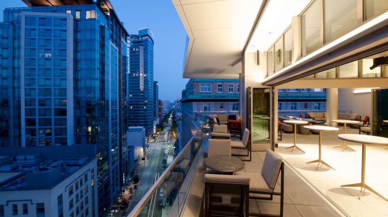 A modern, glass-walled balcony with outdoor seating overlooks a city street surrounded by tall buildings at dusk.