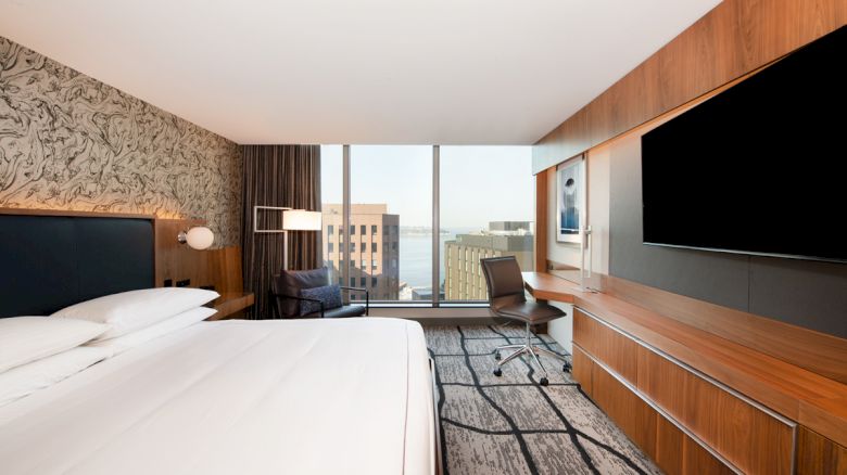The image shows a modern hotel room with a large bed, a flat-screen TV, a desk with a chair, and a view of buildings through a large window.