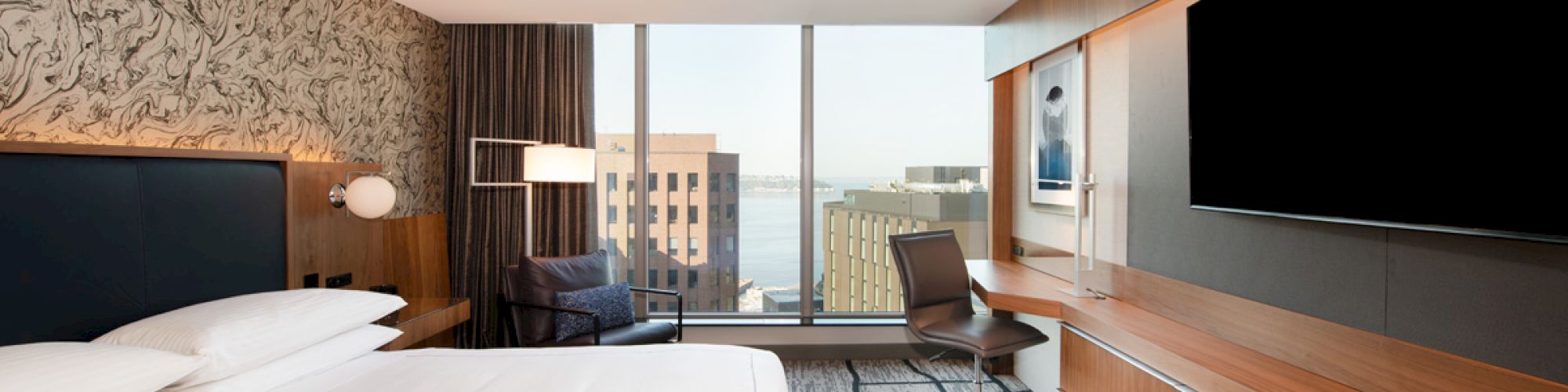 The image shows a modern hotel room with a large bed, desk, chair, wall-mounted TV, and a city view through large windows.