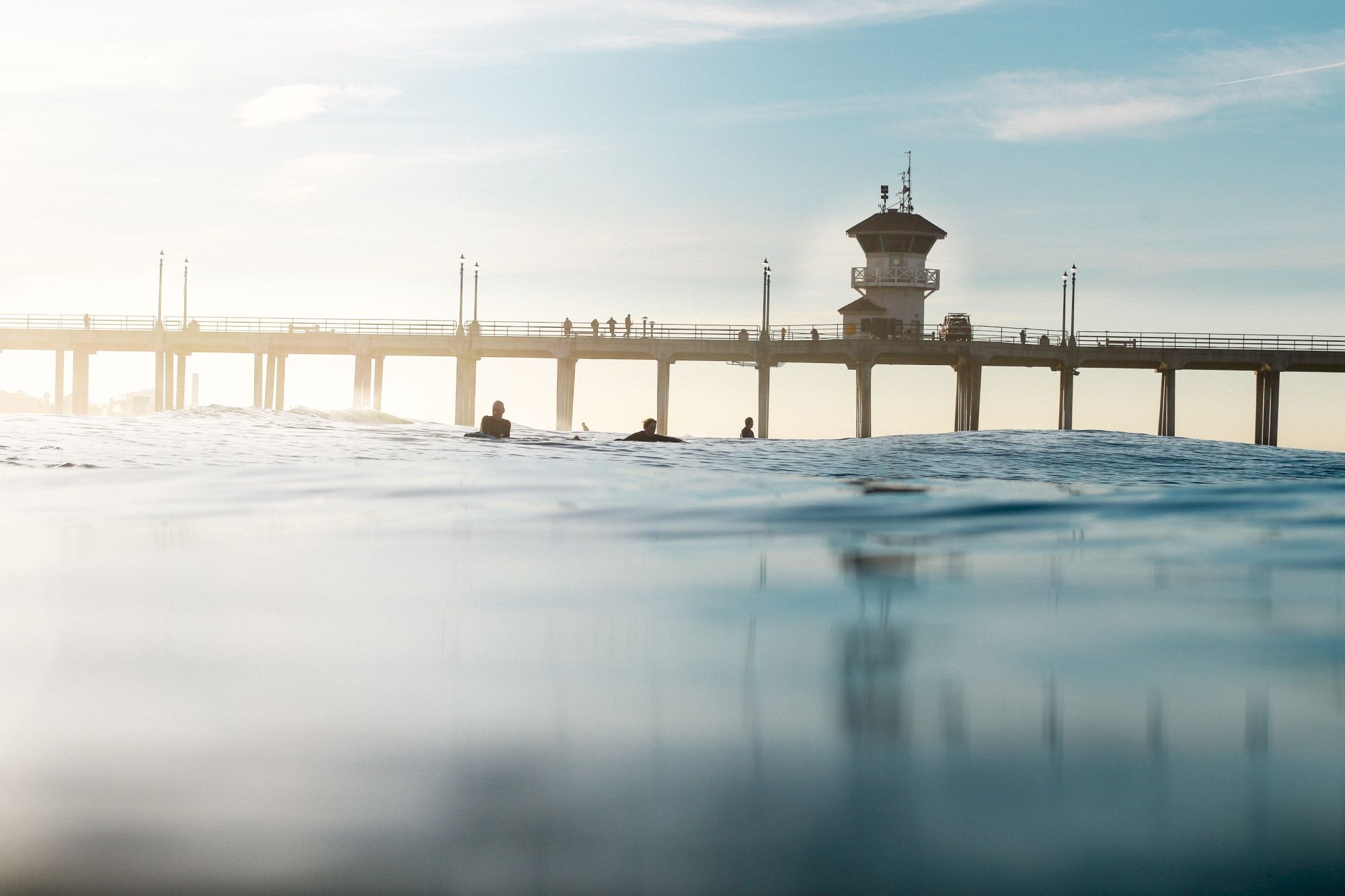 A view of a pier extending over the ocean, with a few surfers in the water and people walking on the pier in the background.
